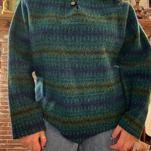 May include: A teal, blue, and olive green patterned sweater with a collared neckline and two gold buttons. The long-sleeved sweater has a textured knit design and a relaxed fit. The garment is paired with blue jeans.