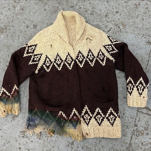 May include: A vintage brown and beige knit cardigan with a geometric pattern. The sweater has a shawl collar, two pockets, and a distressed appearance with holes and frayed edges. The cuffs and hem are also distressed, with faded color accents.