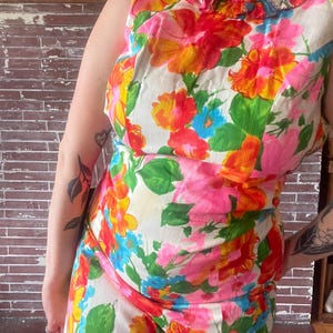May include: A sleeveless floral print dress with a vibrant mix of pink, orange, yellow, and blue flowers on a white background. The dress has a ruffled neckline and a fitted silhouette, showcasing a vintage style.