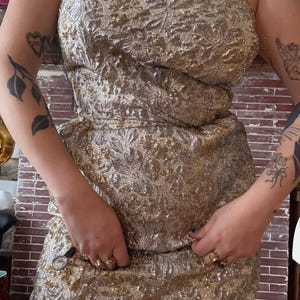 May include: A vintage gold-colored dress with a floral pattern. The dress features a strapless bodice and a fitted waist. The fabric has a textured appearance with metallic threads, creating a shimmering effect. The dress is a formal or evening wear garment.