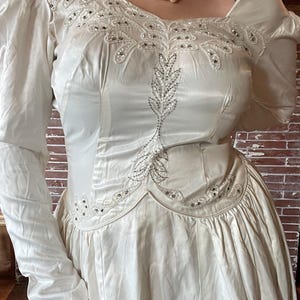 May include: Ivory-colored vintage wedding dress with long sleeves and a square neckline. The bodice features intricate beaded embellishments and a leaf-like design. The dress has a full skirt and a classic, elegant style.