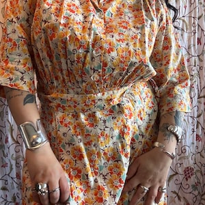 May include: A button-down shirt featuring a vibrant floral pattern in shades of orange, yellow, and teal on a cream background. The shirt has a classic collar, short sleeves, and a matching fabric belt at the waist, creating a structured silhouette.