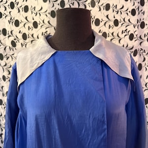 May include: A blue garment with a light grey collar. The fabric has a slight sheen, and the collar is a contrasting colour. The garment is displayed on a mannequin. The background is a floral pattern.
