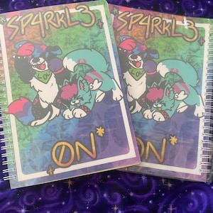 Puede incluir: Dos cuadernos de espiral con cubiertas coloridas que presentan personajes de dibujos animados. Las cubiertas tienen un fondo degradado de arcoíris con el texto "SP4RKL3" y "ON*" impreso en ellas.