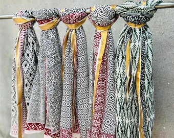 May include: Five white cotton scarves with black and gold geometric patterns. The scarves have gold trim and are hanging from a metal rod.