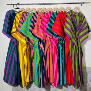 Puede incluir: Una colección de diez caftanes colgados en un perchero de metal, cada uno con rayas verticales vibrantes en varias combinaciones de colores, incluyendo morado y verde, amarillo y naranja, rosa y azul, y rojo y azul verdoso, con cordón.