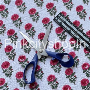 Könnte beinhalten: Weißer Baumwollstoff mit einem sich wiederholenden Muster aus rosa Blüten und grünen Blättern. Eine Schere mit blauen Griffen liegt auf dem Stoff. Ein Lineal ist ebenfalls auf dem Bild zu sehen.