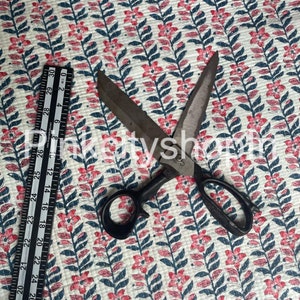 May include: A pair of black handled scissors with silver blades are lying on a white fabric with a repeating pattern of pink flowers and blue leaves. A ruler is visible in the bottom left corner of the image.