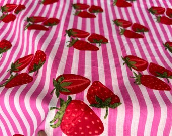 May include: Pink and white striped fabric with a repeating pattern of red strawberries. The strawberries have green stems and leaves. The fabric is folded, showing the pattern's continuity.