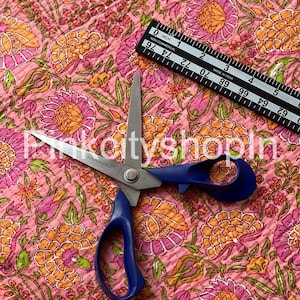 Op de afbeelding: Roze stof met een bloemenpatroon in tinten oranje, roze en groen. Een paar blauwe scharen liggen op de stof. Een zwarte liniaal met witte markeringen ligt ook op de stof. De liniaal geeft "0 inch" en "1 inch" en "Made in India" aan.