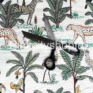 May include: White fabric with a repeating pattern of green palm trees, zebras, leopards, toucans, and birds. A pair of black scissors is placed on top of the fabric.