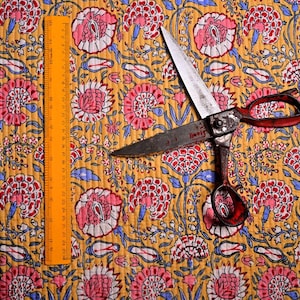 May include: Close-up of floral fabric with a yellow background, featuring pink and red flowers with blue accents. An orange ruler and vintage scissors with red handles are on the fabric.
