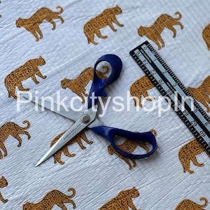 Op de afbeelding: Witte stof met een herhalend patroon van oranje en bruine luipaarden. Een paar blauwe scharen en een zwarte liniaal liggen op de stof.