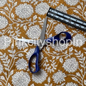 Puede incluir: Primer plano de una tela de color amarillo mostaza con un patrón floral blanco. Un par de tijeras con mangos azules y una regla están sobre la tela.