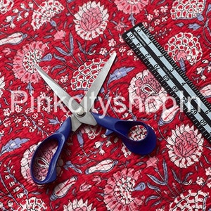 Op de afbeelding: Rode stof met een bloemenprint in roze, wit en blauw. Een schaar met blauwe handvatten ligt op de stof. Een liniaal is gedeeltelijk zichtbaar in de rechterbovenhoek van de afbeelding.