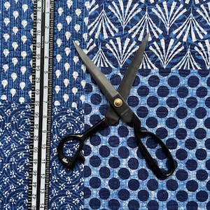 以下が含まれることがあります： 金色のアクセントが付いた黒い布用はさみは、青と白の模様の布の上に置かれています。布には、白い水玉模様、白い花柄、白い幾何学模様の3つの異なる模様があります。