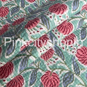 May include: A quilted fabric with a floral pattern. The design features large pink flowers, blue-grey leaves, and white accents on a light teal background. The fabric is stitched with a linear pattern.