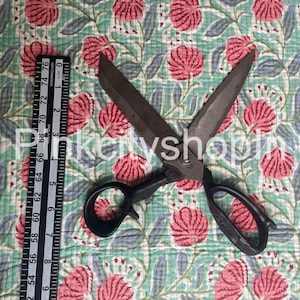 May include: A pair of black handled scissors with a silver blade, lying on a green and pink floral fabric. The fabric has a repeating pattern of flowers and leaves. A ruler is visible in the background.