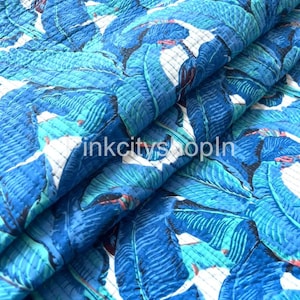 Op de afbeelding: Blauw en groen tropisch bladmotief stof met een gestikte textuur. De stof is dubbelgevouwen en het patroon is aan beide kanten zichtbaar.