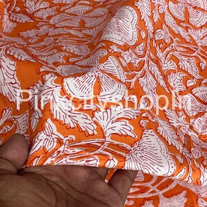 Op de afbeelding: Oranje stof met een witte bloemenprint. De stof heeft een gladde textuur en een subtiele glans.