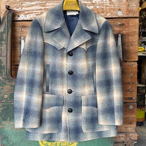 May include: A vintage plaid jacket in shades of blue, beige, and cream. The jacket features a notched collar, four black buttons, and two front pockets. The jacket is hanging on a wooden hanger.