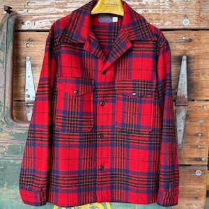 May include: A red and blue plaid flannel shirt with two chest pockets and black buttons. The shirt is hanging on a wooden hanger. The shirt has a classic, vintage style.