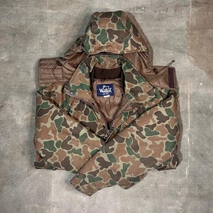 Peut inclure: Une veste à capuche de camouflage marron, vert et beige avec une fermeture éclair. La veste a un patch marron sur la poitrine gauche avec le texte "Woolrich" en blanc.
