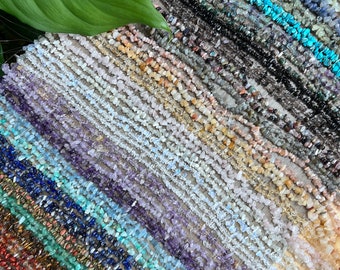 May include: A variety of chip gemstone strands in different colors, including clear, white, pink, purple, blue, green, brown, and black. The strands are laid out in rows on a wooden surface.