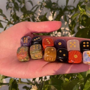 May include: A collection of twelve gemstone dice in various colors, including red, black, and purple, with gold-colored pips. The dice are held in a hand, showcasing their polished surfaces and unique patterns. The dice are for tabletop games.