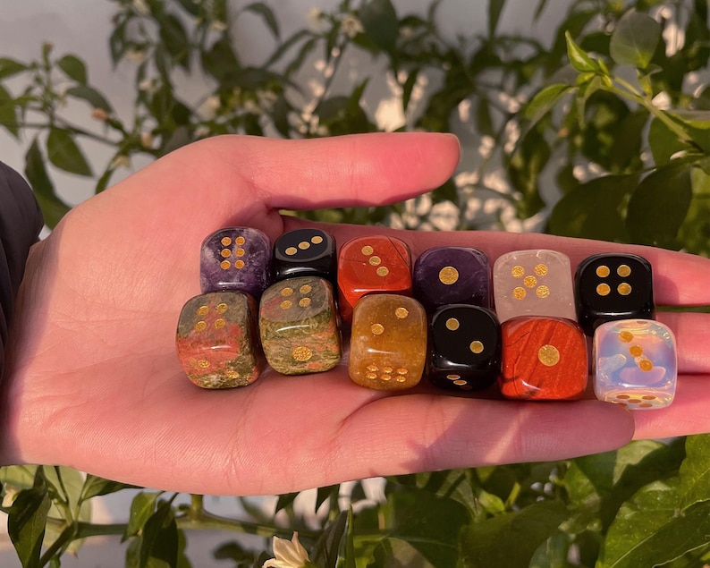 May include: A collection of twelve gemstone dice in various colors, including red, black, and purple, with gold-colored pips. The dice are held in a hand, showcasing their polished surfaces and unique patterns. The dice are for tabletop games.