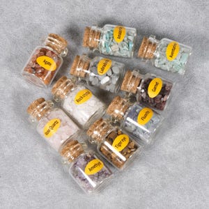 May include: A collection of small glass bottles filled with various gemstones, including agate, amethyst, and rose quartz. Each bottle has a cork stopper and a label with the gemstone's name. The bottles are arranged on a gray surface.