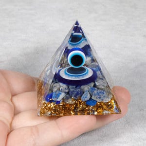 May include: A clear resin pyramid with a blue and white evil eye design. The pyramid is filled with blue stones and gold glitter. The pyramid is held in a hand.