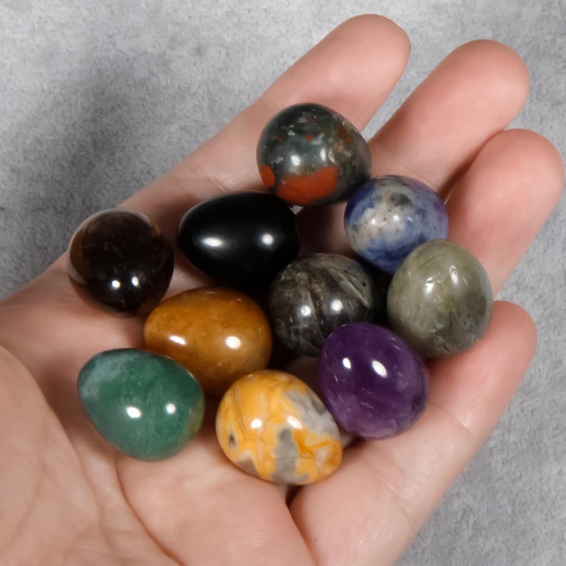Puede incluir: Una colecci&oacute;n de huevos de piedras preciosas pulidas en varios colores, incluyendo verde, morado, amarillo y negro. Los huevos son lisos y brillantes, con patrones y texturas &uacute;nicos. Los huevos se sostienen en una mano.