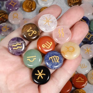 May include: A selection of colourful, round gemstone zodiac symbols in various colours, including purple, green, red, and blue. Each stone features a gold-coloured zodiac symbol. The stones are held in a hand.