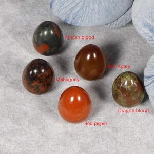Puede incluir: Cinco huevos de piedras preciosas pulidas en tonos rojos, marrones y verdes. Los huevos est&aacute;n etiquetados como "African blood", "Mahagony", "Red Agate", "Red jasper" y "Dragon blood". Las piedras se muestran sobre una superficie gris claro.