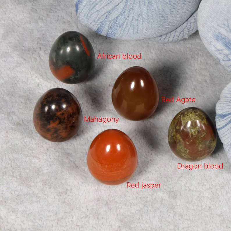Puede incluir: Cinco huevos de piedras preciosas pulidas en tonos rojos, marrones y verdes. Los huevos est&aacute;n etiquetados como "African blood", "Mahagony", "Red Agate", "Red jasper" y "Dragon blood". Las piedras se muestran sobre una superficie gris claro.