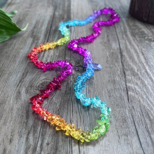 May include: A rainbow-colored necklace made of faceted glass beads. The beads are arranged in a graduated pattern, with the smallest beads at the top and the largest beads at the bottom. The necklace is lying on a wooden surface.