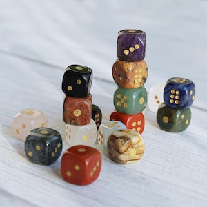 May include: A collection of gemstone dice in various colors, including purple, green, red, and brown. Each die features gold-colored pips. Some dice are stacked, creating small towers. The dice are set against a light, wooden background.