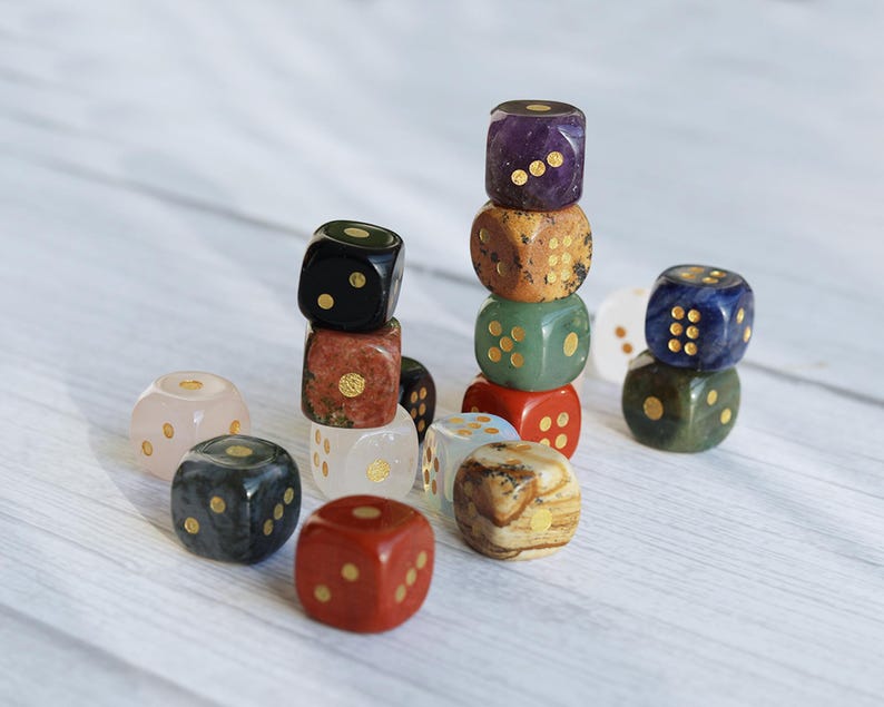 May include: A collection of gemstone dice in various colors, including purple, green, red, and brown. Each die features gold-colored pips. Some dice are stacked, creating small towers. The dice are set against a light, wooden background.
