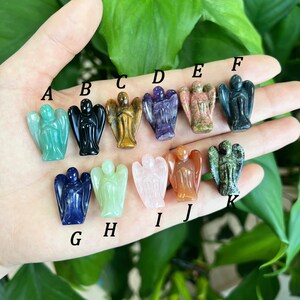 Pode incluir: Uma coleção de 12 pequenas figuras de anjos esculpidas em várias pedras preciosas, incluindo aventurina verde, obsidiana negra, olho de tigre, ametista, quartzo rosa, cornalina e muito mais. As figuras são dispostas em uma fileira em uma mão, com letras que rotulam cada uma.