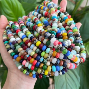 May include: A large collection of colorful gemstone beads, including green, blue, red, yellow, and white beads. The beads are arranged in a circular pattern and are held in a hand.