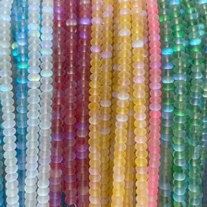 Puede incluir: Múltiples hilos verticales de cuentas de vidrio esmerilado redondas dispuestas en un espectro de colores que incluye blanco, azul, rojo, amarillo, rosa y verde. Cada cuenta presenta un acabado mate suave con un sutil brillo iridiscente.
