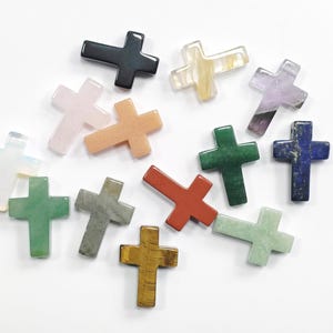 May include: A selection of gemstone crosses in diverse colours, such as green, red, blue, and pink. The crosses are crafted from various materials and have a polished finish. The crosses are arranged on a white surface.
