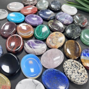 K&ouml;nnte beinhalten: Eine Sammlung glatter, polierter ovaler Steine in verschiedenen Farben und Mustern. Die Steine sind in einem Halbkreis auf einer dunklen Oberfl&auml;che angeordnet. Einige der Steine umfassen Amethyst, Rosenquarz, Tigerauge und Jaspis.