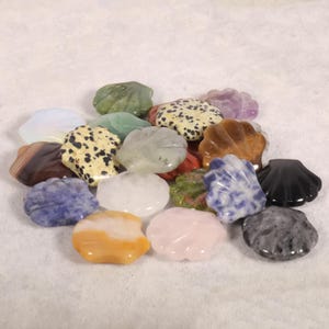 May include: A collection of various gemstone shell carvings in shades of green, yellow, pink, brown, black, and blue. The polished stones have unique patterns and textures, showcasing natural variations. The shells are arranged on a white surface.