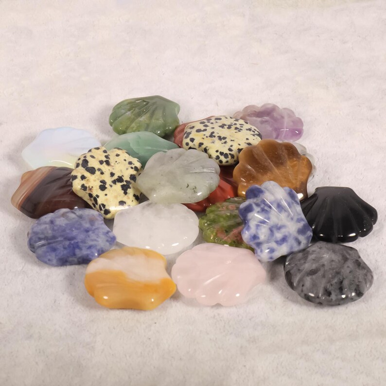 May include: A collection of various gemstone shell carvings in shades of green, yellow, pink, brown, black, and blue. The polished stones have unique patterns and textures, showcasing natural variations. The shells are arranged on a white surface.