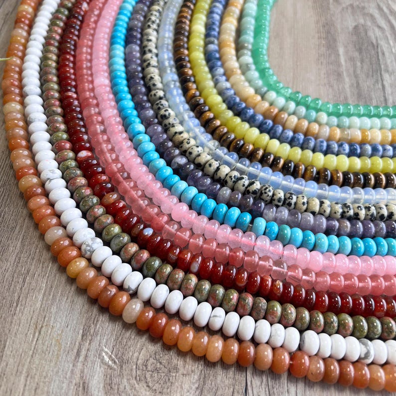May include: A variety of gemstone beads in different colors and patterns, including red, orange, yellow, green, blue, purple, and white. The beads are arranged in a semi-circle on a wooden surface.