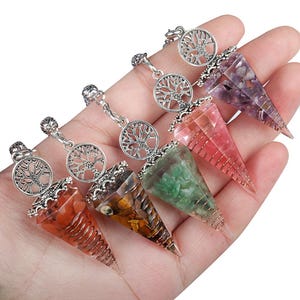 Puede incluir: Una colección de péndulos de cristal en varios colores, incluyendo rojo, verde, morado y marrón. Cada péndulo tiene un dije plateado con un árbol de la vida y una forma de cono envuelta en alambre. Los péndulos se sostienen en una mano.