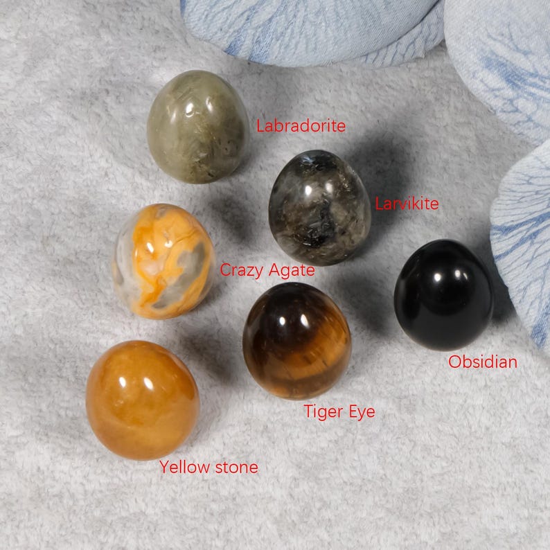 Puede incluir: Un surtido de huevos de piedras preciosas pulidas, incluyendo Labradorita, Larvikita, Crazy Agate, Ojo de Tigre, Piedra Amarilla y Obsidiana. Los huevos var&iacute;an en color, desde amarillo y naranja hasta negro y gris, sobre una superficie gris claro.