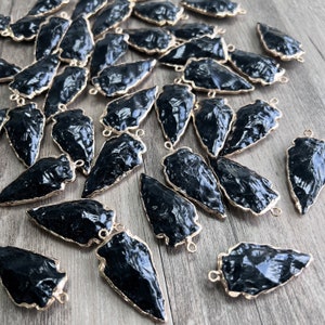 May include: A collection of black arrowhead-shaped pendants with gold-toned metal loops. The arrowheads are made of a smooth, black material and have a slightly textured surface.